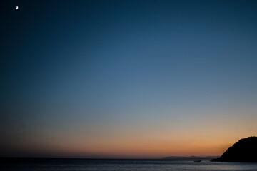 Fototapeta premium A mysterious dusk unfolds as the moon rises over the tranquil sea, casting an enchanting glow on the coastal waters in the serene embrace of twilight.