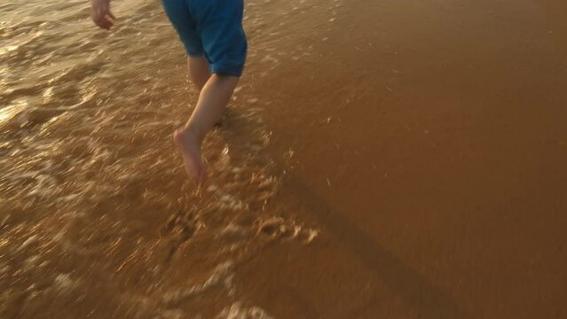 Back view of kid running barefoot along ocean coast at sunset.