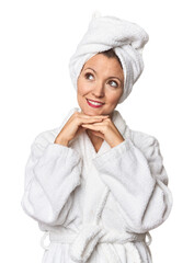 Caucasian woman in bathrobe and towel keeps hands under chin, is looking happily aside.