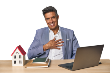 Middle-aged Hispanic businessman with real estate theme laughs out loudly keeping hand on chest.