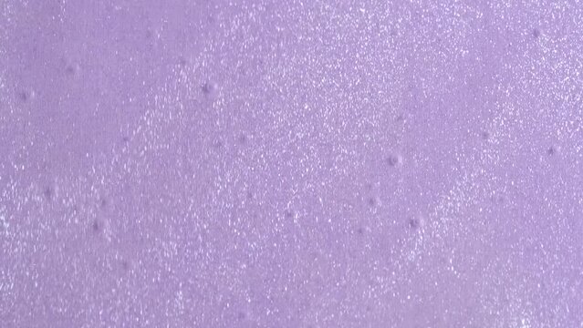 Shimmering vivid silver liquid glowing particles flow motion on lavender paper background. Glowing particles. Transparent glittering shower gel or shiny lotion filling the screen.