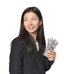 Young Chinese woman with dollars in suit looks aside smiling, cheerful and pleasant.