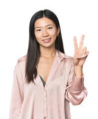 Young Chinese woman in studio setting joyful and carefree showing a peace symbol with fingers.