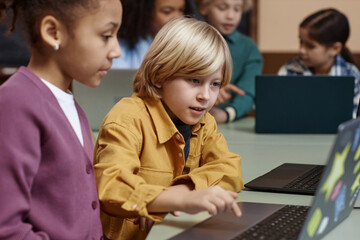Side view portrait of blonde little boy using laptop and doing online tests in school classroom