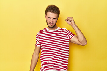 Young Caucasian man on a yellow studio background showing fist to camera, aggressive facial...