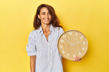 Middle aged woman holding a wall clock on a yellow backdrop happy, smiling and cheerful.