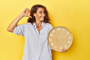 Middle aged woman holding a wall clock on a yellow backdrop feels proud and self confident, example...