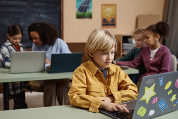 Fototapeta premium Portrait of blonde young boy using laptop computer in school classroom, copy space