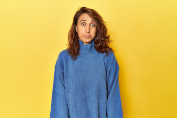 Fototapeta premium Middle-aged woman on a yellow backdrop blows cheeks, has tired expression. Facial expression concept.