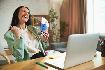Happy, joyful young woman, employee sitting in office with laptop, holding plane tickets and...