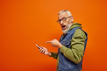 surprised handsome mature man in green hoodie pointing at smartphone and looking at camera