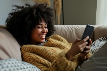 Mixed race woman smiling while scrolling social media on her smartphone, cozy vibes in the sofa at home. 
