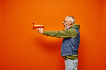 handsome mature male model in green hoodie and stylish denim vest aiming with megaphone in hands