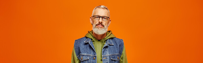 funky mature man in vibrant hoodie and stylish vest looking at camera on orange backdrop, banner