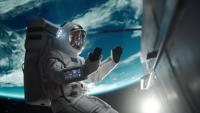 Young Astronaut Floating in Space in Zero Gravity. Spaceman Using a Smartphone in Open Space Outside an Orbiting Spacecraft, Having a Video Call with Launch Operations Center on Earth