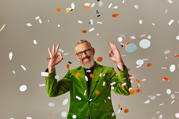 handsome jolly mature man in green blazer showing okay sign under confetti rain on beige backdrop