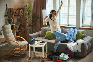 Happy and excited young woman at home, listening to music in headphones, dancing and packing her suitcase for travelling. Concept of traveling, tourism, vacation, emotions
