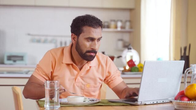 Indian Middle Aged Man Working On Laptop While Eating Food On Dining Table At Home - Concept Of Modern Lifestyle, Work From Home And Surfing Web