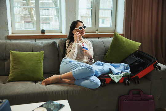 Happy, joyful young woman sitting at home, holding plane tickets, talking on phone and expressing excitement about upcoming trip. Concept of traveling, tourism, vacation, emotions