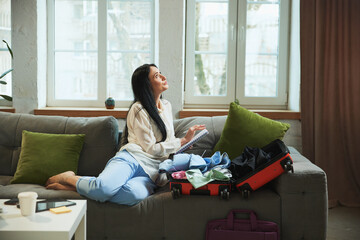 Happy and excitement young woman sitting on sofa at home with suitcase packed, writing notes and...