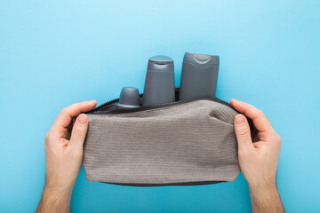 Young man hands holding gray cosmetic travel bag with plastic bottles on pastel blue table...