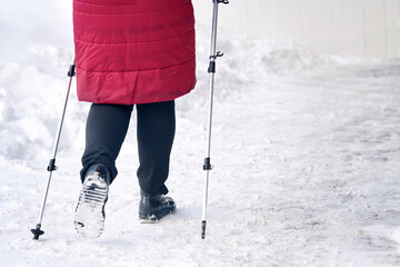 Senior woman walking along slippery snowy sidewalk with walking poles. Old woman practicing Nordic...
