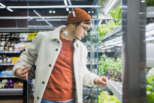A man in the supermarket chooses fresh microgreens for a salad, healthy vegetarian food rich in vitamins and fiber. Adult male in a warm coat selecting vegetables in a grocery store. 