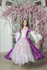 Obraz na płótnie Canvas Beautiful woman in fantasy white and purple rococo style medieval dress dancing near door with pink flowers