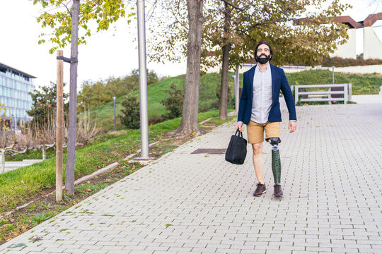 Confident man with a prosthetic leg and a stylish outfit walking on a cobbled path in a serene park environment.