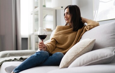 Portrait of pretty, charming, attractive, stylish woman sitting on couch having raised glass with red wine in hand, examine, taste beverage.