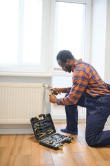 Man in workwear overalls using tools while installing or repairing heating radiator in room