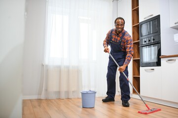 Young african man washes the floor with a mop in the room - obrazy, fototapety, plakaty