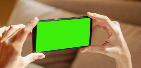 Close up of hands of lying at couch woman holding smartphone with green screen on blurred backdrop.