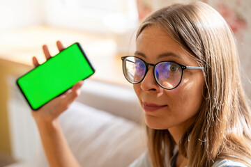 Young girl in eyeglasses with long hair holds smartphone with green screen in hand looking at camera