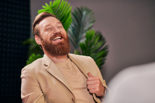 handsome cheerful man with beard in elegant attire laughing during interview while in studio