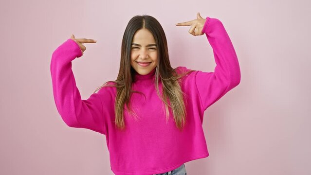 Confident young latin woman in pink background, smiling proudly and pointing at self with finger in her sweater standing tall. advertisement for self-esteem and success with a pinch of silliness