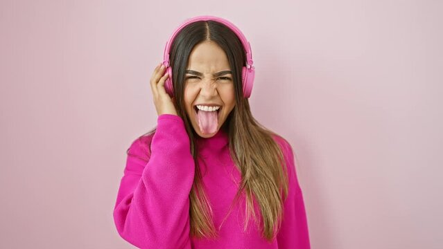 Beautiful young latin woman sticking out tongue in fun expression while listening to music, happy and amusing over pink isolated background