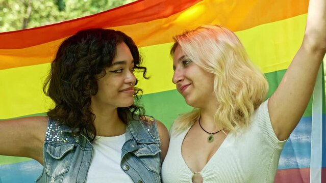 two young lesbian girls in slow motion holding a gay pride flag above them kissing