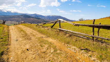 country road through the hill with grassy meadow behind the wooden fence. carpathian mountain ridge with snow capped tops in the distance. beauty of ukrainian countryside in spring