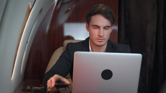 Rich successful businessman flies on private jet and works. Businessman uses laptop on business class plane while sitting on window seat surrounded by gadgets. Luxurious lifestyle of businessman.