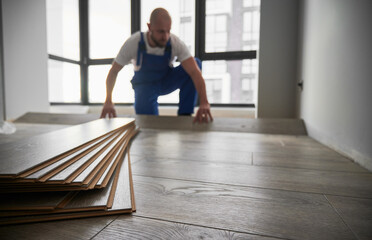 Selective focus of laminate wooden planks and male construction worker on blurred background. Mane installing laminate timber floor in apartment under renovation.