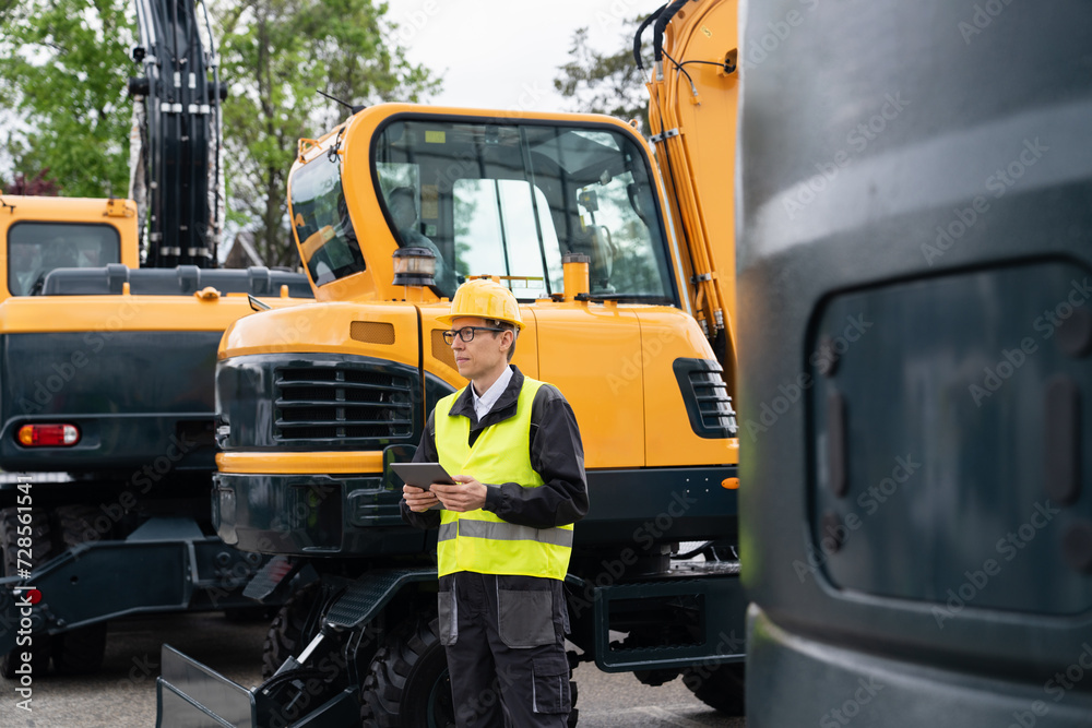 Poster Engineer in a helmet with a digital tablet stands next to construction excavators..