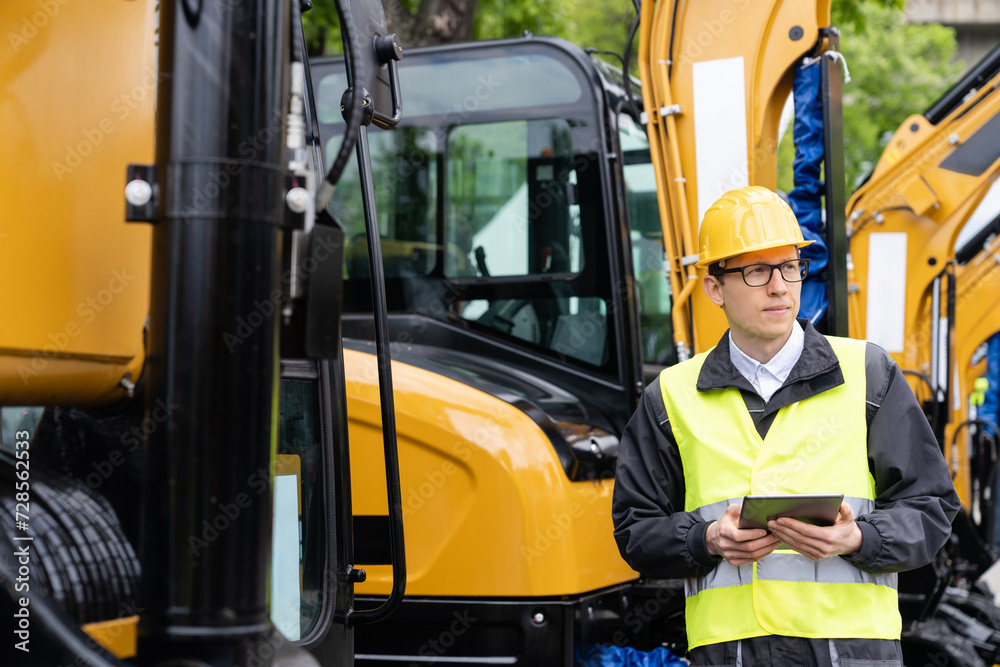 Poster engineer in a helmet with a digital tablet stands next to construction excavators..