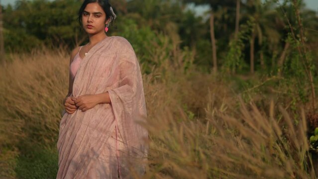 Woman in pink saree standing contemplatively in golden field, soft focus, dusk light
