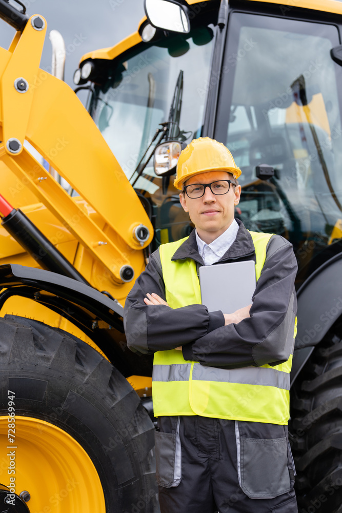 Poster engineer in a helmet with a digital tablet stands next to construction excavators..