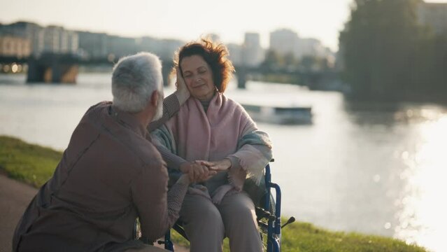 Husband walking with wife in wheelchair. Senior family couple spending time in city park near pond. Man helping female showing love care, they embrace. Living with physical disability concept.