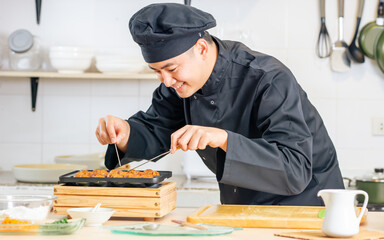 Portrait close up handsome professional Japanese male chef wearing black uniform, hat, cooking, making takoyaki street food, smiling with confidence, standing in kitchen. Restaurant, Hotel Concept.