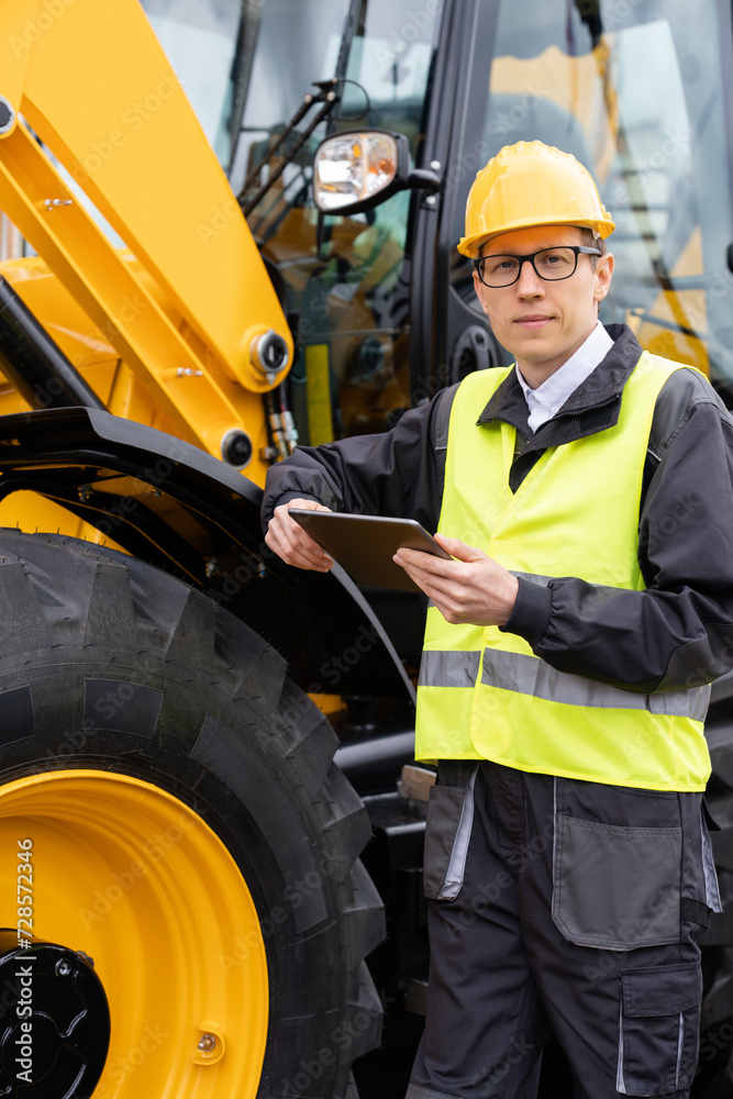 Poster Engineer in a helmet with a digital tablet stands next to construction excavators..