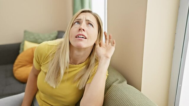 Attractive young blonde woman at home, hand over ear, intently listening and hearing gossip or rumor - deafness concept. happy portrait in t-shirt.