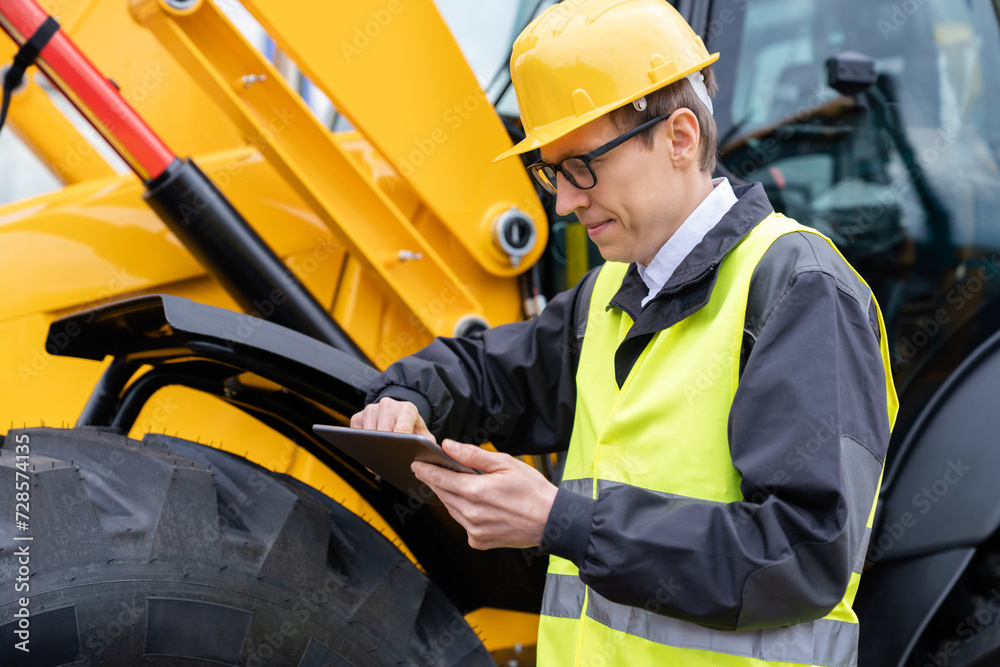 Poster Engineer in a helmet with a digital tablet stands next to construction excavators..